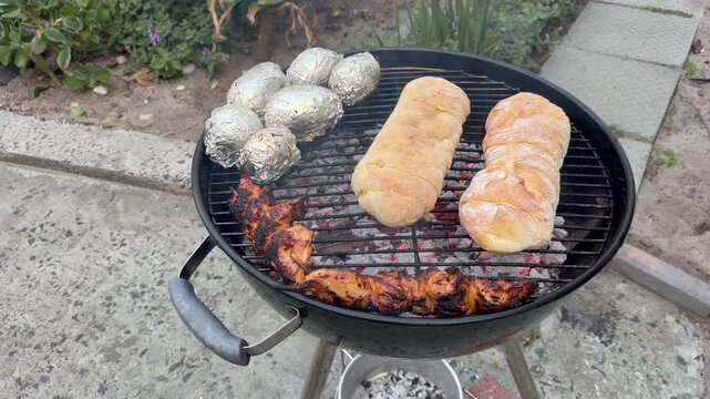 A round charcoal braai grill loaded with charred grilled chicken, golden bread rolls toasting, and foil-wrapped potatoes roasting over glowing coals, set outdoors on a paved patio.