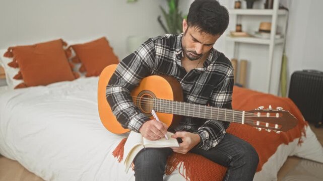 Man with classical guitar writing notes by hand on a notebook while seated on a bed in a building; concentration songwriting focus.