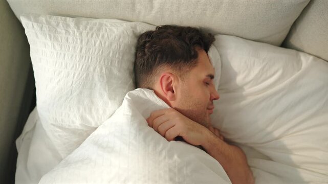 A young man sleeps on his side clutching a white duvet, then rolls onto his back and smiles with eyes closed. Top view of a cozy bed with seersucker pillows, beige sofa cushions and soft morning sun.