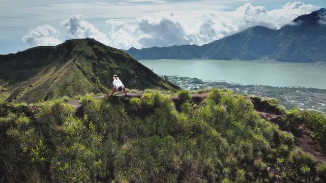 Athlete man holds a planche calisthenics pose on a mountain ridge above Lake Batur, Bali, showcasing strength, balance, and dedication against a volcanic landscape and blue sky. Aerial drone panorama