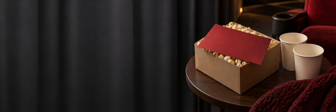 Popcorn in a box with paper cups beside cinema seats. Movie night snack setup for film viewing and home entertainment. Cinema concession treat. Advertising banner with ample copy space