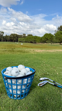 Basket of used golf balls and golf clubs on green grass at an outdoor driving range with spacious golf course and blue cloudy sky in background.