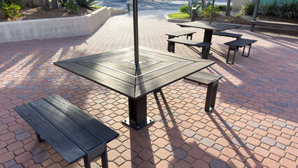 dark outdoor picnic tables and benches sit on a reddish brick paved patio under bright afternoon sunlight © DrewDuzz