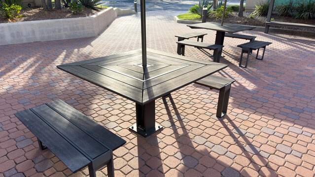 dark outdoor picnic tables and benches sit on a reddish brick paved patio under bright afternoon sunlight