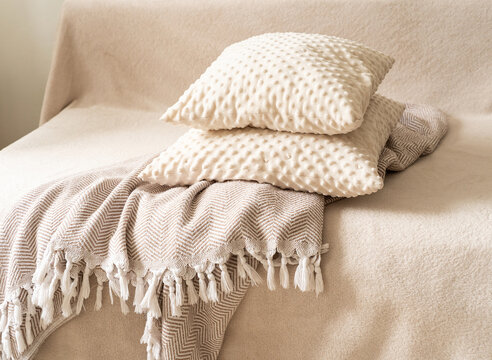 Textured decorative pillows in milky-beige hue, stacked atop patterned throw blanket with white fringe, resting on beige sofa against neutral backdrop. Quiet luxury, cozy home accessories