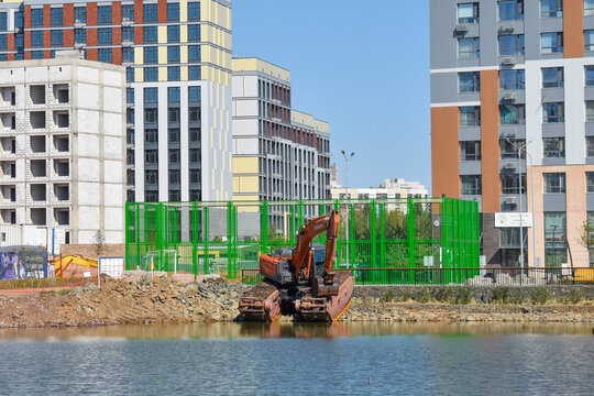 Construction machinery and an excavator on the shore of a water body against the background of a modern residential complex construction and site landscaping