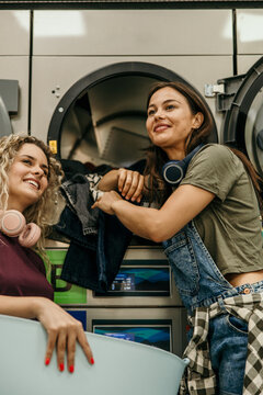 Young women friends doing laundry together smiling in laundromat