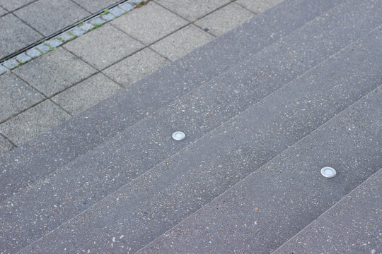 Close-up view of gray concrete stairs with embedded round lights and textured surface, adjacent to patterned paving stones in an outdoor urban setting
