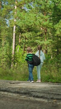 Boy and girl carry black backpacks as they stroll along paved trail in woods. scene features tall green trees with sunlight filtering through leaves grassy undergrowth on both sides of path
