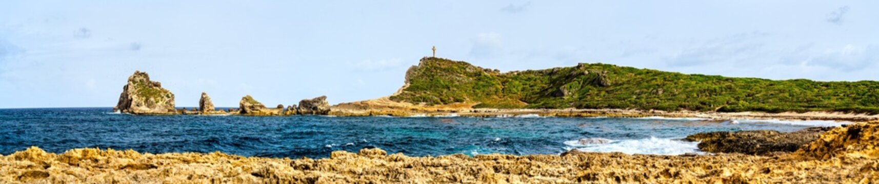 Wide panoramic view of the rocky coastline, turquoise ocean waters, and hills with the summit cross at Pointe des Chateaux in Guadeloupe, Caribbean France