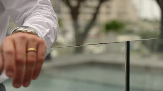 A close-up shows a person in a white shirt handling the top edge of a glass pool gate, with a wristwatch and ring visible against a softly blurred background.