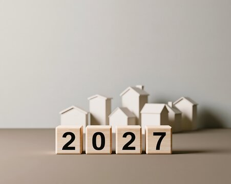Four wooden blocks displaying the year 2027 in front of a collection of small, white, minimalist house models on a grey surface