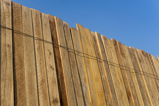 Rough sawn wooden planks arranged vertically outdoors against a clear blue sky, showing natural wood grain, uneven edges and weathered surfaces in warm sunlight.