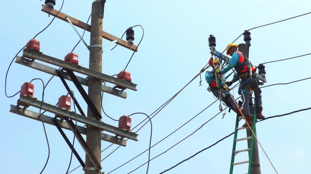 Two electricians are working at height on a three-phase power line. They are wearing helmets and safety gear. They handle the cables and insulators with care.