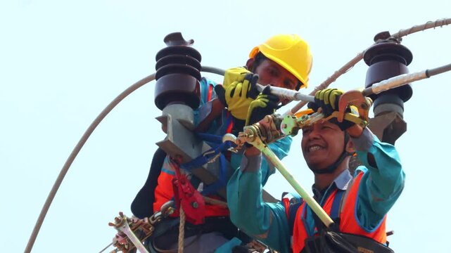 Two electricians work high on power lines. They wear helmets and safety gear. They manage wires and insulators with care. Their teamwork shows skilled, focused effort