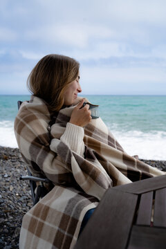 A young cozy woman is sitting by the seaside, wrapped in a warm beige and brown blanket, enjoying quiet solitude in the cool day's loneliness with a warm drink.