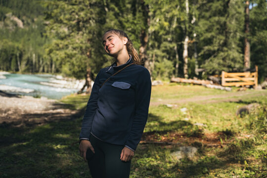 A young woman is relaxing in outdoor clothing in a wooded setting surrounded by trees and sunlight, during an outdoor journey. Active travel and relaxation concepts.