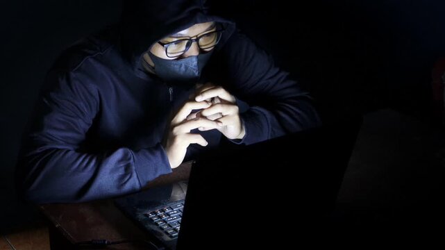 Male hacker wearing a hood, glasses, and face mask while working on a laptop in a dark room, accessing an online website or security system.