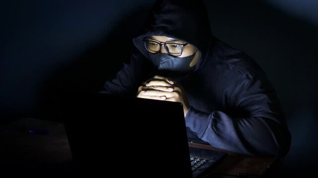 Male hacker wearing a hood, glasses, and face mask while working on a laptop in a dark room, accessing an online website or security system.