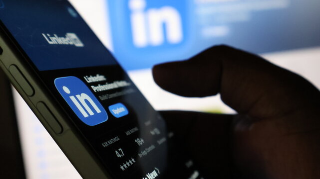 Person holding smartphone displaying LinkedIn mobile application store interface in front of computer monitor showing brand logo.