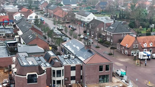 Wide cinematic parallax shot of a building undergoing renovation in the center of a Dutch village, featuring newly installed solar panels on the roof, with surrounding houses and village streets visib