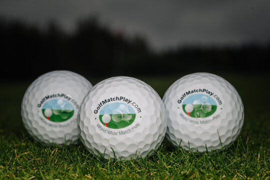 three golfmatchplay.com golf balls arranged on green grass with forest and cloudy sky beyond, detailed dimples and printed logos in a close up sports product scene