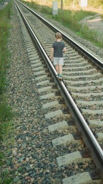 Small child carefully balances and moves forward on parallel train tracks. scene is set in outdoor environment featuring gravel paths alongside greenery. lighting suggests bright sunny day