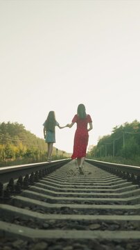 Two young women hold hands as they stroll along parallel railroad tracks. One wears red dress with white patterns and other is dressed casually both move away from camera into open landscape