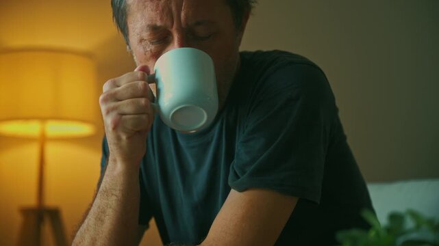 A contemplative middle-aged man drinking from a coffee mug in warm living room light, appearing thoughtful and alone as he quietly reflects during an evening at home. 4K.
