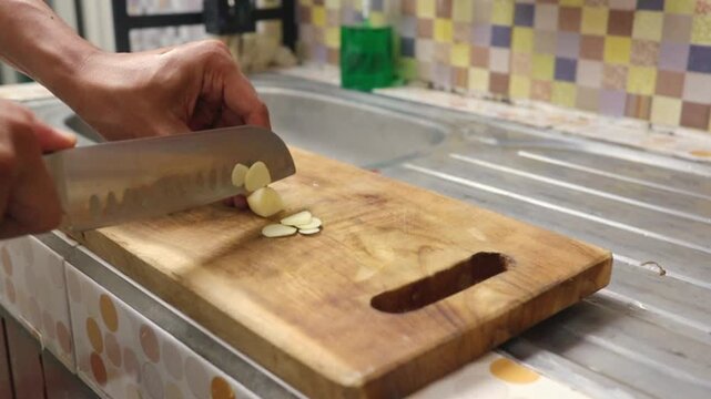 man hands slicing fresh garlic cloves using a sharp knife on a wooden cutting board in a home kitchen for cooking