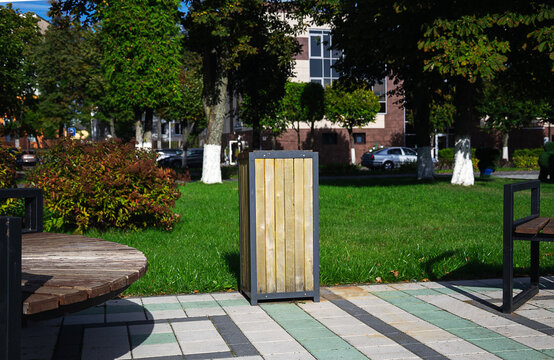 Wooden waste bin standing between park benches on tiled pavement of public square with green lawns and trees on sunny day