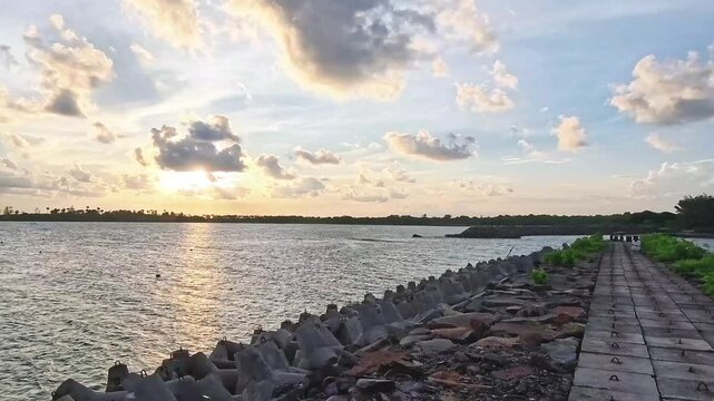 Glowing sunlight bursting through scattered clouds over calm sea waters, bordered by concrete breakwater tetrapods along the coast.