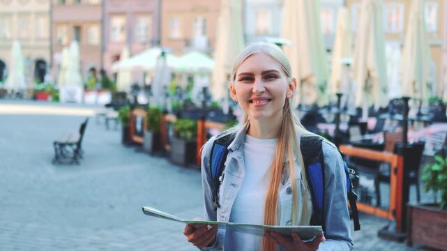 A young blonde woman holding a folded paper map and smiling at the camera in a European urban outdoor market plaza with cafe seating and classical architecture.