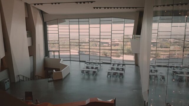 Still high-angle view of empty tables beneath tall windows in a concrete interior.