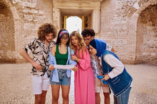 Diverse Group Of Young Friends Studying Map While Exploring Historic Stone Archway On City Walking Tour