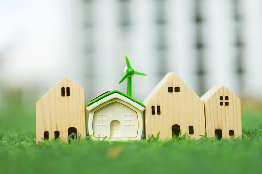 Green energy eco house and sustainable living concept, model house with solar roof and wind turbine among wooden houses on lawn, representing renewable real estate, and clean architecture.