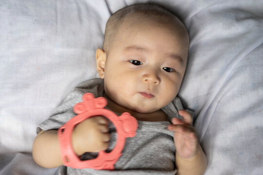 Infant teething and baby sensory development concept, cute Asian baby boy in grey clothes lying down and holding pink silicone teether ring, representing pediatric oral health, and childcare.