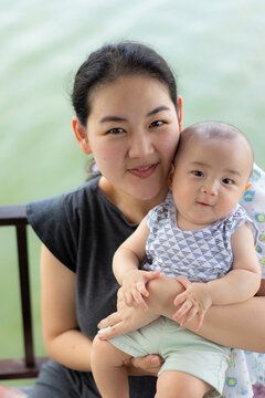 Motherhood love and family bonding concept, happy smiling young Asian mother holding cute chubby baby in arms outdoors by balcony railing, representing maternal healthcare, and parenting joy.
