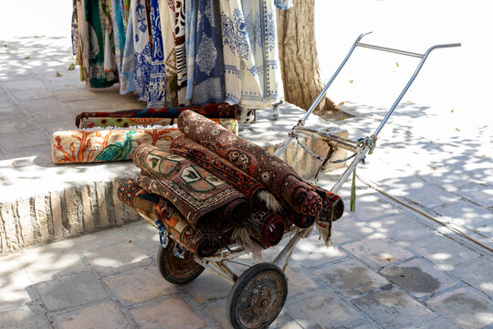Bukhara, Uzbekistan. Rolled patterned rugs rest on a two wheeled handcart in the foreground, while colorful textiles hang behind them beside a tree on a sunlit paved walkway