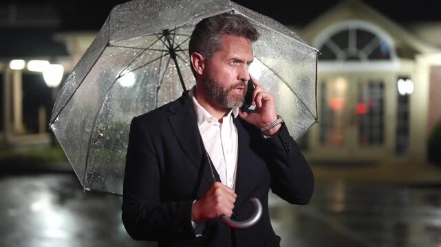 Businessman talks on his phone under an umbrella on a rainy night. Manager makes a business call in the rain. Man in suit speaking on a mobile phone in night street. Business man checks his schedule.