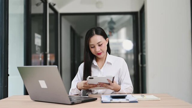Smiling Asian businesswoman multitasking with a digital tablet and laptop at a modern office desk. A confident professional working online