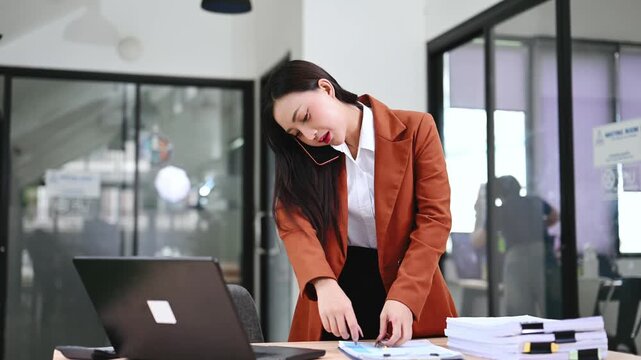 Smiling Asian businesswoman multitasking at her desk, talking on a smartphone and using a laptop in a modern office.