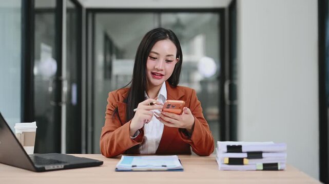 Smiling Asian businesswoman multitasking with a digital tablet and laptop at a modern office desk. A confident professional working online