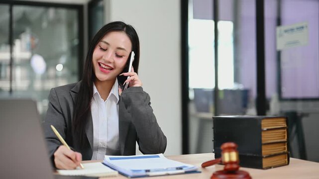 Asian female attorney multitasking on a call while reviewing contracts. A portrait of professional success at a desk with a gavel and scales of justice.
