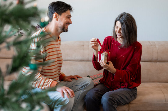Mature man watches woman opening Christmas gift on sofa