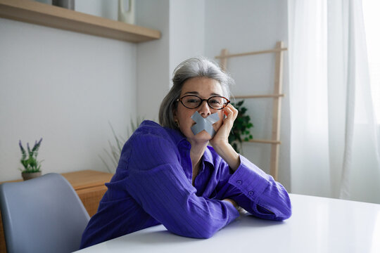 Mature woman with tape over mouth sitting at table indoors representing censorship