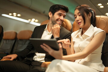Two seatmates sharing a tablet share a tablet screen in adjacent seats in the passenger cabin of a plane, both clearly enjoying the conversation, good for connection and travel themes © ultramansk