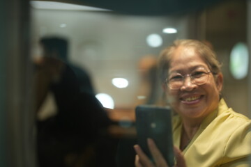 An older Asian traveller wearing glasses relaxes in her aircraft seat with her phone in the cabin of a passenger aircraft, good for retirement and travel themes © ultramansk