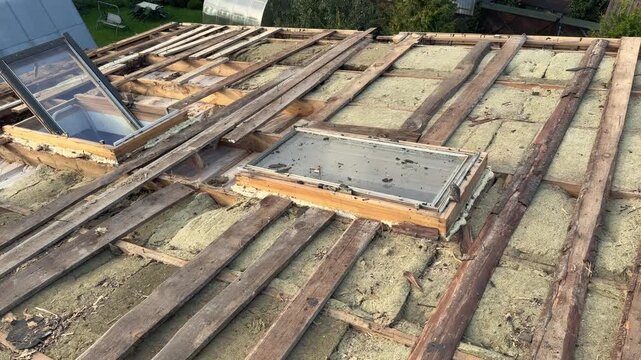 Exposed wooden roof framing, aged insulation, and skylights are visible as a residential roof undergoes repair and renovation work