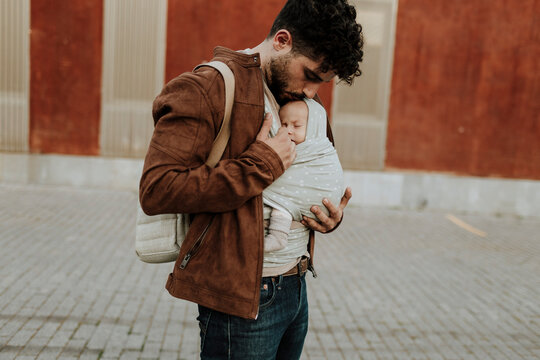 A father kisses his baby in a carrier on an urban street